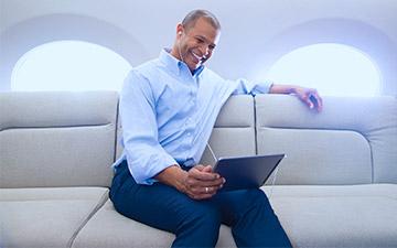 微笑的男人坐在私人飞机上，使用私人飞机互联网, 穿着蓝色纽扣衬衫看着平板电脑