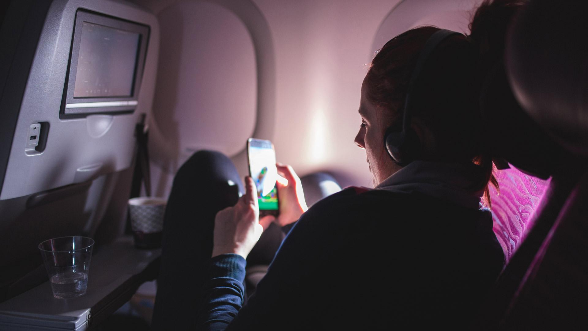 A person with headphones on curls up in an airplane seat and looks at something on a mobile phone screen. 